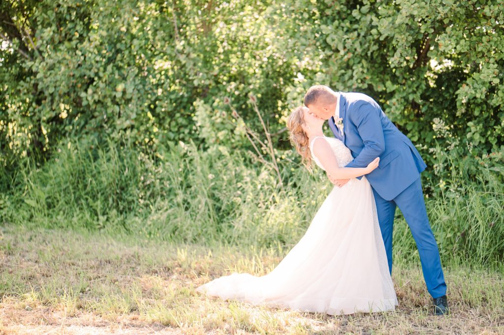 Aiden Laurette Photography | brunette man in blue suit and blonde in wedding dress kiss in front of trees