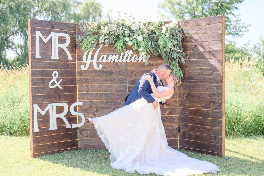 Aiden Laurette Photography | bride and groom kiss in front of wooden backdrop that reads "MR & MRS HAMILTON"